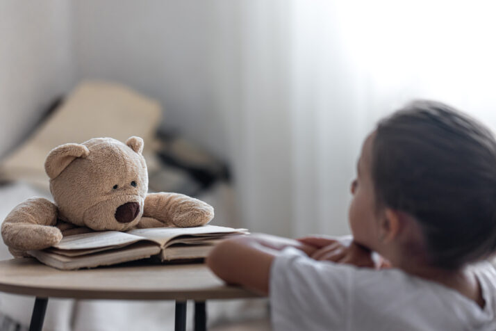 A little girl plays with her teddy bear and a book, teaches him to read, plays at school.