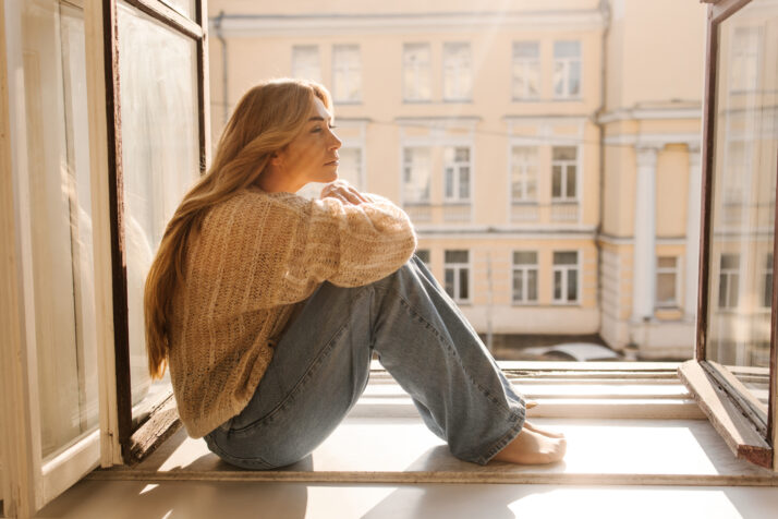 Sad fair-skinned adult woman blonde in warm sweater and jeans, sitting on windowsill on sunny day. Mood concept