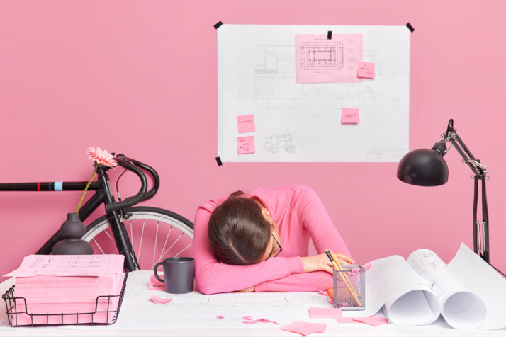 Exhausted tired overworked female engineer works all day on new design project leans at table wants to sleep surrounded with sketches and blueprints poses in home office. Lack of productivity