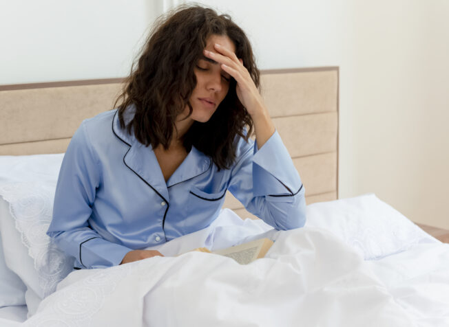 funny young beautiful woman in blue pajamas sitting in bed with book touching head feeling bad suffering from headache in bedroom interior on light background